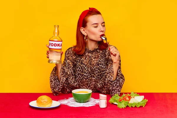 一个时髦的红头发女人在黄色的背景下坐在桌旁吃饭 吃着汤 喝着伏特加 食品流行艺术摄影 互补的颜色 文字的复制空间 — 图库照片
