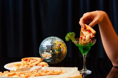 Parti zamanı. Yemek ve sanat fotoğrafçılığı. Siyah stüdyo arka planıyla pizza dilimini kokteyle koyan erkek eli yakın plan görüntüsü. Yaratıcılık. Tamamlayıcı renkler. İlan, metin için boşluk kopyala