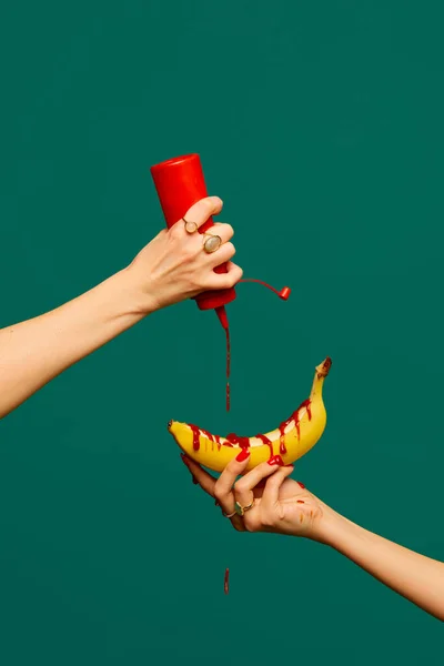 Female hand pouring ketchup at banana against green studio. Extraordinary taste. Food pop art photography. Creativity, art. Complementary colors. Copy space for ad, text