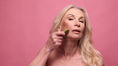 Sağlıklı cilt masajı yapan orta yaşlı, güzel bir kadın pembe stüdyo arka planında gua-sha ile. Doğal güzellik kavramı, yüz bakımı, kozmetoloji ve kozmetik, sağlık