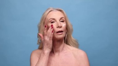 Güzel orta yaşlı kadın nemlendirici yüz kremi sürüyor. Mavi stüdyo geçmişine karşı poz veren olgun yaşlı bir kadın. Doğal güzellik kavramı, yüz bakımı, kozmetoloji ve kozmetik, sağlık