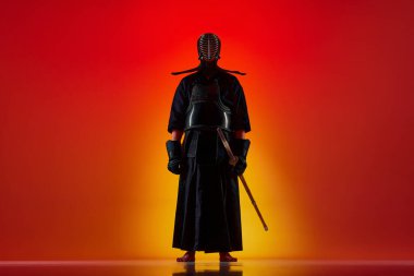 İnsan portresi, üniformalı profesyonel kendo atleti Shinai kılıcıyla neon ışıklarıyla gradyan kırmızı stüdyo arka planına poz veriyor. Dövüş sanatları, spor, Japon kültürü, eylem, hareket
