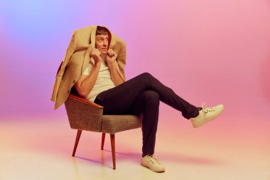 Olgun, orta yaşlı, poz veren, sandalyede oturan ve parlak ışıkta pembe arkaplanda ceketin altına saklanan bir adam portresi. Duyguların kavramı, yaşam tarzı, yüz ifadesi, eğlence. Ad