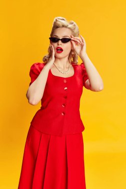 Şok olmuş yüzüyle sarı stüdyo arka planına poz veren kırmızı takım elbiseli, şık saç stili ve modaya uygun güneş gözlüklü güzel bir genç kız. Eski moda anlayışı, güzellik, 50 'ler, 60' lar. Pin-up tarzı