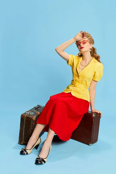 感觉很疲倦穿着黄色衬衫和红色裙子的年轻漂亮的女孩坐在蓝色工作室背景的老式手提箱上 复古时尚的概念 小鸡鸡风格 — 图库照片