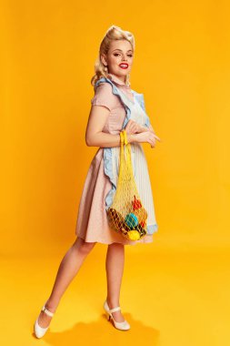 Market alışverişi. Sarı stüdyo arka planında net çantasıyla poz veren şık saç stili güzel bir genç kızın portresi. Eski moda anlayışı, güzellik, 50 'ler, 60' lar. Pin-up tarzı