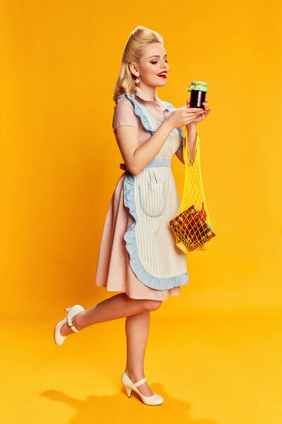 Food shopping. Portrait of beautiful young girl with stylish hairstyle posing against yellow studio background. Concept of retro fashion, beauty, 50s, 60s. Pin-up style