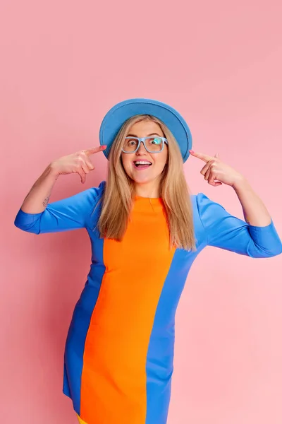Positive emotions. Portrait of woman in colorful clothes, hat and glasses emotionally posing against pink studio background. Concept of emotions, facial expression, lifestyle and fashion