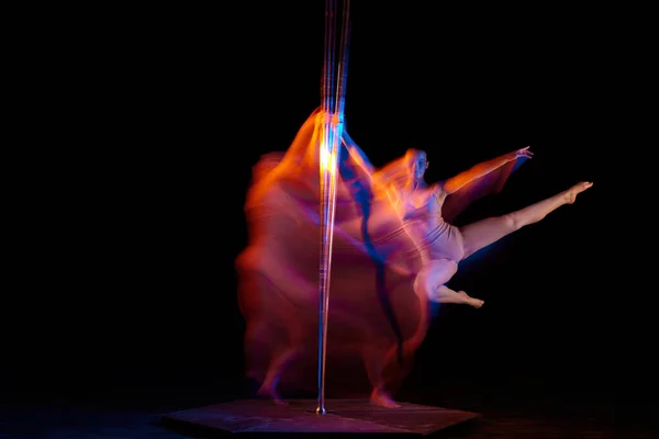 In dynamics, spinning. Young girl performing pole dance isolated over black studio background with mixed neon lights. Concept of sport and dance, beauty of movements, action, modern style