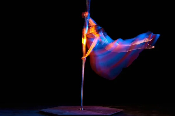 Strong hands. Young girl dancing, performing pole dance isolated over black studio background with mixed neon lights. Concept of sport and dance, beauty of movements, action, modern style