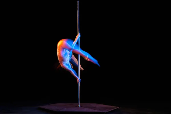 Doing tricks. Young girl performing pole dance isolated over black studio background with mixed neon lights. Concept of sport and dance, beauty of movements, action, modern style