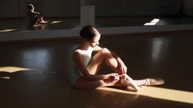 Tender teen girl, classical ballet dancer getting ready to train in ballet school on a daytime with sunlight. Putting on pointe shoes. Concept of ballet, dance art, education, beauty, choreography