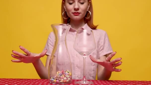 フードポップアート写真 黄色のスタジオの背景に複数の色のキャンディーをワイングラスに注ぐスタイリッシュな若い女性 ヴィンテージでレトロなスタイル 補完的な色 テキストのコピースペース — ストック動画