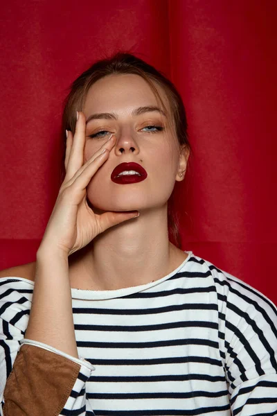 Simple stylish look. Portrait of young beautiful girl with red lips and hair bun, in striped shirt posing against red studio background. Concept of female beauty, fashion, modern style, youth