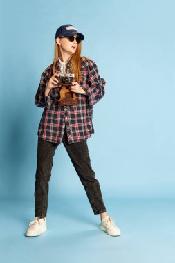 Traveller. Young beautiful girl in checkered shirt, cap and sunglasses posing with photo camera on blue studio background. Concept of youth, beauty, fashion, lifestyle, emotions, facial expression. Ad clipart