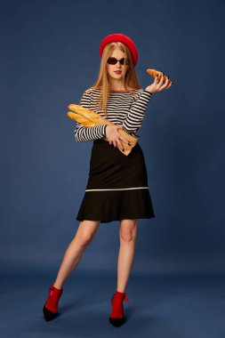 Kırmızı bereli güzel bir kızın portresi. Mavi stüdyo arka planında baget ve kruvasanla poz veriyor. Gençlik kavramı, güzellik, moda, yaşam tarzı, duygular, yüz ifadesi. Ad