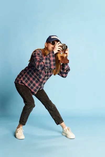 摄影师 穿着格子衬衫 帽子和太阳镜的年轻漂亮的女孩在蓝色的工作室背景上摆出照相相机的姿势 生活方式 面部表情的概念 — 图库照片