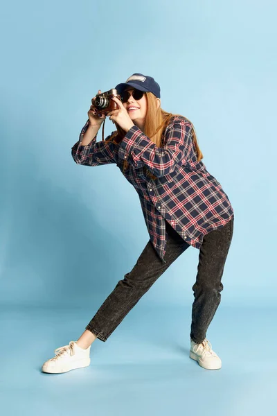 穿着格子衬衫 帽子和太阳镜的年轻漂亮的女孩在蓝色的工作室背景上摆出照相相机的姿势 生活方式 面部表情的概念 — 图库照片