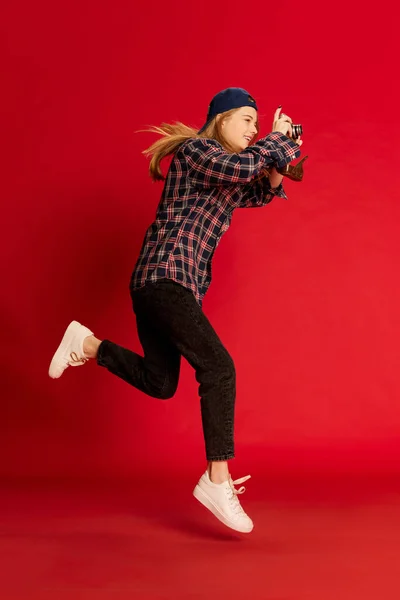 穿着格子衬衫 帽子和太阳镜的年轻貌美的女人跳过红色的摄影棚背景拍照 生活方式 面部表情的概念 — 图库照片