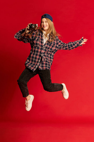 Красивая молодая женщина в клетчатой рубашке, кепке и солнцезащитных очках фотографируется с камерой на фоне красной студии. Концепция молодости, красоты, моды, образа жизни, эмоций, выражения лица. Объявление