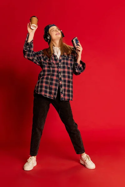 Energy. Young beautiful positive girl in checkered shirt and cap listening to music in headphones, dancing over red studio background. Concept of youth, fashion, lifestyle, emotions, facial expression