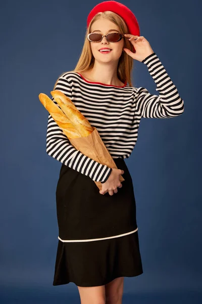 穿着红色贝雷帽的年轻漂亮姑娘的画像 与蓝色工作室背景下的法式法式面包合影 巴黎风格 生活方式 面部表情的概念 — 图库照片
