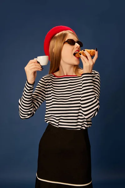 穿着红色贝雷帽的漂亮女孩的画像 在蓝色工作室背景下吃着带有咖啡的羊角面包 生活方式 面部表情的概念 — 图库照片
