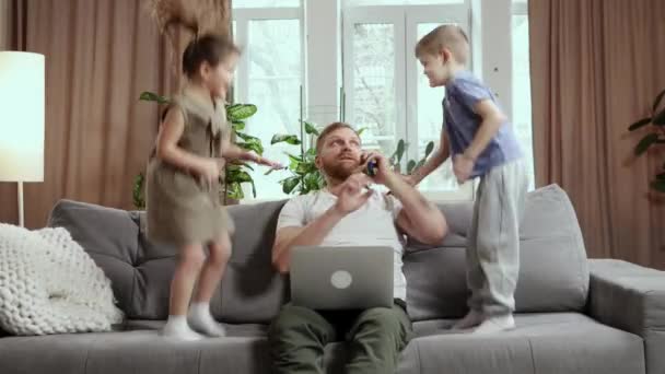 ビジネスマン 父はリビングルームでソファに座って 仕事をして電話で話しています 男の子と女の子 子供たちが飛び回って彼を妨害する 子供時代 フリーランス リモートワークの概念 — ストック動画