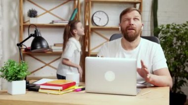 Evde çalışan sakallı, olgun bir adam, online iş toplantısı, konferans. Küçük kız onun etrafında yüksek sesle oynuyor. Babalık kavramı, çocukluk, serbest meslek, uzak çalışma, duygular