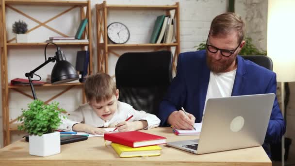 留着胡子的男人 父亲坐在桌旁 带着他的小儿子画画 在家里的笔记本电脑上工作 家庭聚会 自由职业 远程工作 情感的概念 — 图库视频影像
