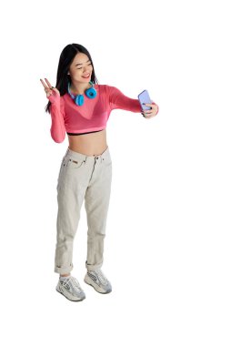 Üst Manzara. Günlük kıyafetlerle gülümseyen, cep telefonuyla selfie çeken, beyaz arka planda görüntülü görüşme yapan genç bir kız. İş anlayışı, eğitim. Yaşam tarzı. İlan için alanı kopyala