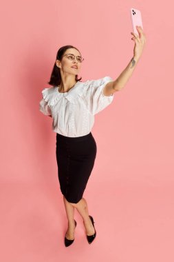 Resmi giyinmiş, cep telefonlu selfie çeken güzel bir kadının resmi. Pembe stüdyo arka planında. İş anlayışı, eğitim, meslek, duygular, moda. Ad