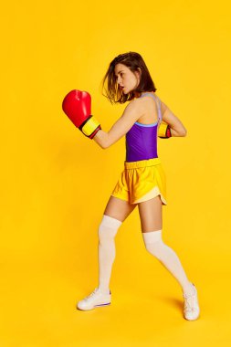 Kırmızı boks eldivenleriyle parlak sarı stüdyo arka planına poz veren şık spor elbiseli genç bir kadının tam boy görüntüsü. Spor anlayışı, sağlıklı yaşam tarzı, duygular, moda, güzellik. Ad
