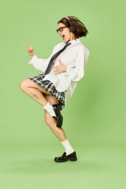 Kulaklıkla müzik dinleyen duygusal genç bir kadının tam boy görüntüsü yeşil stüdyonun arka planında. İllüzyon gitarı. Gençlik kavramı, moda, duygular, yaşam tarzı, yüz ifadesi. Ad