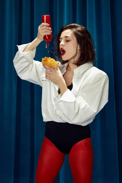 Junk food lover. Putting ketchup on fries. Young beautiful brunette woman in white shirt and red tights posing against blue textile background. Concept of beauty, fashion, emotions, lifestyle. Ad