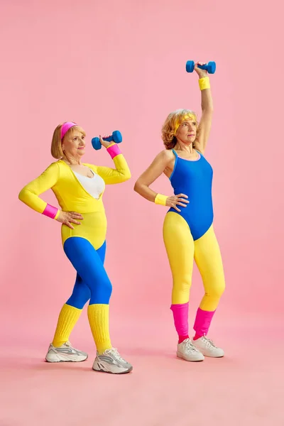 ピンクのスタジオの背景にダンベルを持ち上げ カラフルなスポーツウェアのトレーニングで2人のシニア中年のスポーツ女性 スポーツライフスタイル ウェルネスの概念 — ストック写真