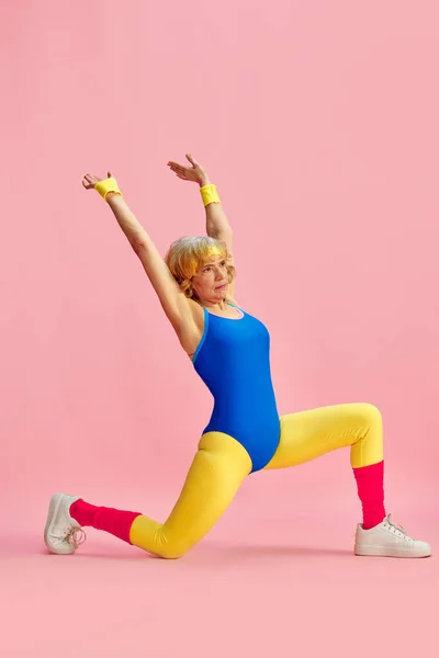 Flexible sportive elderly woman in colorful sportswear training, doing stretching exercises against pink studio background. Concept of sportive lifestyle, retirement, health care, wellness. Ad