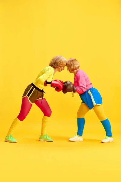 Round. Two elderly sportive woman in colorful uniform training, boxing, posing against yellow studio background. Concept of sportive lifestyle, retirement, health care, wellness. Ad