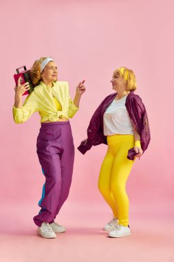 İki orta yaşlı, yaşlı, renkli üniformalı, radyo dinleyen ve dans eden, pembe stüdyo geçmişine karşı poz veren kadın. Sportif yaşam tarzı, emeklilik, sağlık, sağlık. Ad