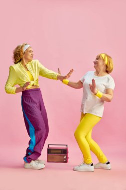 İki orta yaşlı, yaşlı, renkli üniformalı, radyo dinleyen ve dans eden, pembe stüdyo geçmişine karşı poz veren kadın. Sportif yaşam tarzı, emeklilik, sağlık, sağlık. Ad