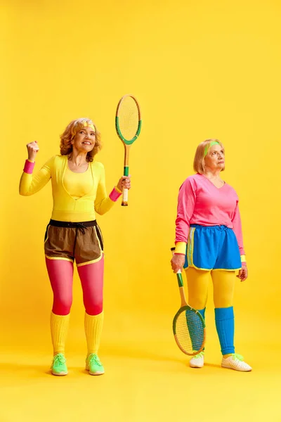 两名中年妇女 穿着五颜六色的制服 穿着网球拍 在黄色的工作室背景下进行训练 运动生活方式 医疗保健 健康的概念 — 图库照片