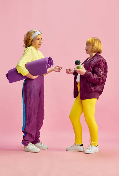 Friends meeting. Elderly sportive woman in colorful uniform posing with fitness equipment against pink studio background. Concept of sportive lifestyle, retirement, health care, wellness. Ad