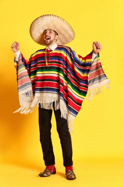 黄色の背景に対してポーズをカラフルなお祝いの服 ポンチョやソムブレロで幸せな興奮顔を持つ若いハンサムな男の肖像画 メキシコの伝統 お祝い 感情の概念 — ストック写真