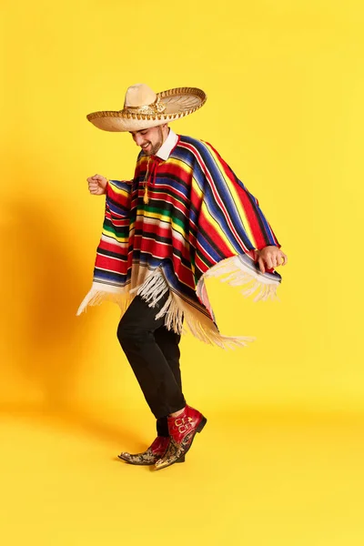 カラフルなお祝いの服 ポンチョやいくつかのポーズで若いハンサムな男の肖像画は 黄色のスタジオの背景に対して踊ります メキシコの伝統 楽しさ お祝い 感情の概念 — ストック写真