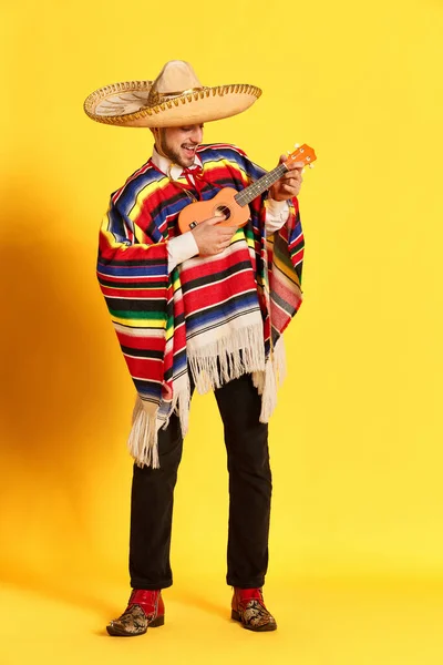 カラフルな服 ポンチョや黄色のスタジオの背景に対してポーズをギターを演奏いくつかのブレロで若いハンサムな男の肖像画 メキシコの伝統 楽しさ お祝い 感情の概念 — ストック写真