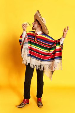 Renkli giysiler içinde genç bir adamın portresi, panço ve sombrero pozu, sarı stüdyo arka planında taco yemek. Meksika gelenekleri kavramı, eğlence, kutlama, festival, duygular