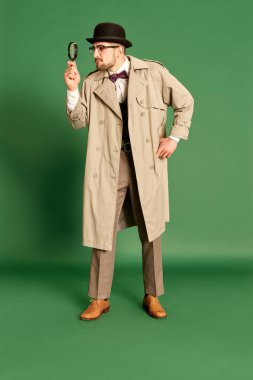 Yeşil stüdyo arka planında büyüteçle bakan şık paltolu ve şapkalı yakışıklı bir gencin tam boy portresi. Duygular kavramı, dedektiflik mesleği, sırlar, moda