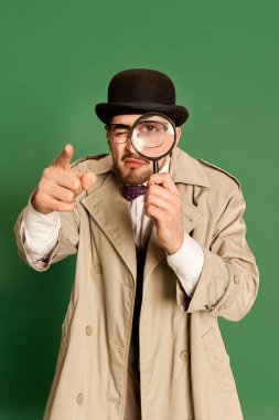Detektif suretinde genç bir adam, şık bir palto ve şapka giymiş, büyüteçle yeşil stüdyo arka planına bakıyor. Duygu anlayışı, dedektiflik mesleği, sırlar, moda. Ad