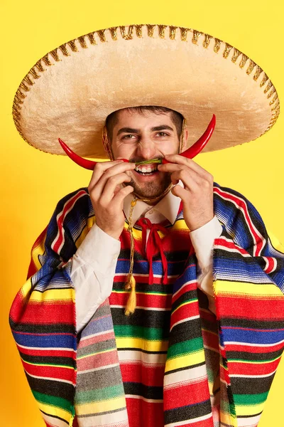 カラフルなお祝いの服を着た若い男 ポンチョと黄色のスタジオの背景に口ひげのような唐辛子でポーズいくつかのブレロ メキシコの伝統 お祝い 感情の概念 — ストック写真