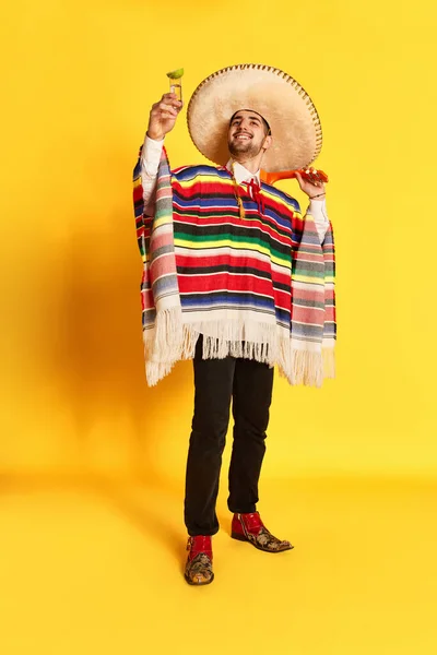 カラフルなお祝いの服を着た若い笑顔の男 ポンチョと黄色のスタジオの背景にテキーラとライムでポーズを取るいくつかのブレロ メキシコの伝統 お祝い 感情の概念 — ストック写真